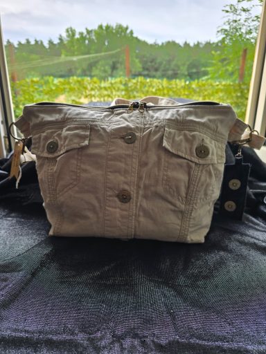 Beige Tasche mit zwei vorderen Taschen und Knöpfen, auf einem dunklen Stoff platziert.
