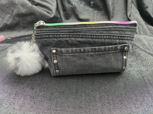 Grauartige Kosmetiktasche mit buntem Reißverschluss und einem weißen Pompon.