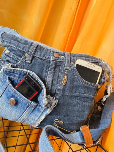 Tasche aus Jeans mit kleineren Taschen und Smartphone darin, vor orangefarbenem Hintergrund.